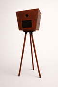 De Luxo Photobooth van Luxo is een cabine in vintage stijl met een houten rechthoekige doos, drie driepoten, een kleine ronde lens en een rechthoekig scherm - ideaal voor bruiloften of evenementen. Weergegeven tegen een effen witte achtergrond.